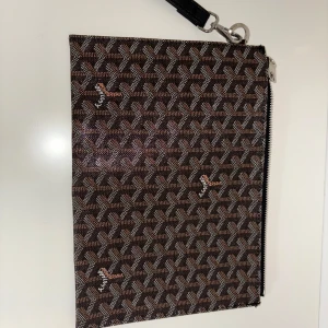 Brun kuvertväska från  - Snygg kuvertväska  i brunt med ikoniskt grafiskt mönster och silverfärgad dragkedja. Väskan har en avtagbar handledsrem och är perfekt för att bära det viktigaste. Klassisk och stilren design som sticker ut.