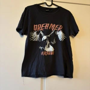 Svart t-shirt med tryck Dreamer - Cool svart t-shirt med tryck av en örn och texten 'Dreamer' och 'Arizona' i röd och vit färg. Klassisk rund hals och korta ärmar. Perfekt för dig som gillar streetstyle och grafiska motiv.