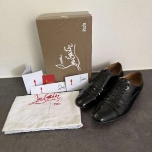 Svarta lackskor från Christian Louboutin - Snygga svarta lackskor från Christian Louboutin med klassisk spetsig tå och ikonisk röd sula. Skorna har snörning och är tillverkade i glansigt skinn med läderfoder. Perfekta för dig som vill sticka ut med exklusiv stil och detaljer.