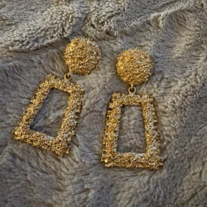 Glittriga guldiga statement örhängen - Säljer ett par stora, glittriga örhängen i guld med rektangulär form och rund topp. De är täckta av små gnistrande stenar som ger en riktigt lyxig och iögonfallande look. Perfekta för dig som vill sticka ut och älskar bling!