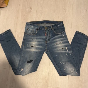Dsquared2 blå slitna jeans, stl 46 - Snygga blå jeans från Dsquared2 med coola slitningar, färgstänk och patch baktill. Jeansen har klassisk femficksdesign, normal passform och är tillverkade i bomull. Perfekta för dig som gillar en edgy streetstil.