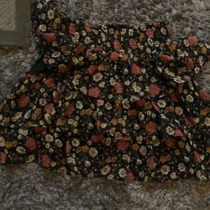 Blommig volangkjol från Lindex - Supersöt kort volangkjol från Lindex med svart botten och blommigt mönster i rosa, gul och beige. Kjolen har flera lager volanger och resår i midjan för en bekväm passform. Perfekt för dig som gillar färg och mönster!