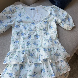 Blommig kortklänning från Design by Amor - Supersöt kortklänning med blått blommönster på vit botten från Design by Amor. Klänningen har v-ringning, puffärmar, spetsdetaljer och volanger i flera lager. Perfekt för sommarens alla tillfällen! Fick hem den idag en tyvärr passade den inte. Priset kan diskuteras vid snabba och seriösa köp