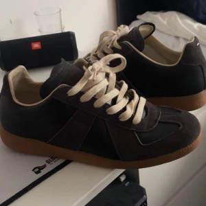 Maison margiela - Snygga svarta  sneakers från Maison Margiela med beige snörning och bruna gummisulor. Skorna har en mix av skinn och mocka, klassisk rund tå och retrovibbar. Perfekta för dig som gillar stilrena och tidlösa sneakers med en twist. Jag kan även tvätta skorna innan dom skickas