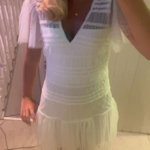 Vit kortklänning med volang och spets - Superfin vit kortklänning med v-ringning, volangkant. Klänningen har spetsdetaljer och omlottkänsla upptill, samt dragkedja i ryggen. Perfekt för sommarens alla tillfällen.