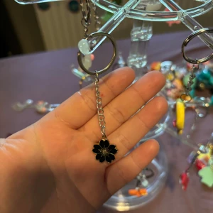 Silverfärgat nyckelring med svart blomma - Snyggt örhänge i silverfärg med en hängande svart blomma i emalj och gulddetaljer. Blomman har en stilren design och örhänget har en tunn kedja som ger en cool och trendig look.