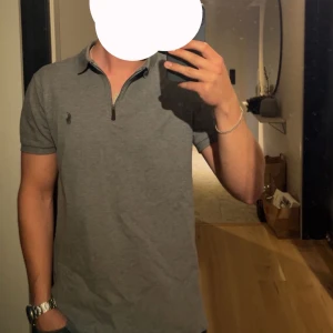 Grå pikétröja Polo Ralph Lauren - Snygg grå pikétröja från Polo Ralph Lauren med klassisk krage och half zip-dragkedja. Tröjan har korta ärmar och den ikoniska broderade loggan på bröstet. Perfekt för en clean och stilren look.