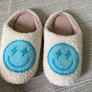 Vita fluffiga tofflor med smiley - Mysiga beiga tofflor i teddy-material med stora blå smiley-märken framtill. Perfekta att glida runt i hemma när du vill ha det extra bekvämt och varmt om fötterna.