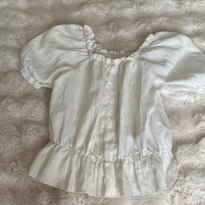 Vit blus med puffärmar och volang - Supersöt vit blus med puffiga korta ärmar, volangkant nertill och knappar framtill. Blusen har en lätt rynkad struktur i tyget och en luftig känsla, perfekt för varma dagar. Rundad halsringning med resår och diskret rutmönster i väven.