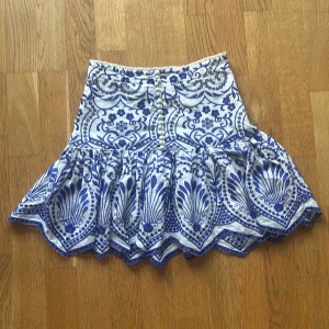 Vit kjol med blått mönster från Zara  - Supersöt kort vit kjol från Zara med blåa broderade blommor och dekorativa knappar framtill. Kjolen har volangkant och spetsdetalj upptill perfekt till dig som vill ha en söt kjol till sommaren. Kjolen knäpps med en dragkedja och är i 100% bomull. Storlek xs men tajt i midjan. Bra skick men säljs pågrund av att den är för liten 💙
