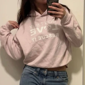 Ljusrosa hoodie HundarUtanHem - Ljusrosa hoodie med vit text framtill: 'DON'T BUY LOVE RESCUE IT' och HundarUtanHem-tryck på ryggen. Klassisk huva, ribbade muddar och normal passform. Perfekt för dig som vill visa stöd för hundar och gillar en chill stil.