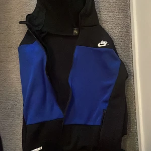 Nike Air blå och svart kofta - Nike Air kofta  i svart och blått med vit logga på bröstet och ärmen. Jackan har huva med dragsko och dragkedja framtill. Sportig look och perfekt för dig som gillar streetwear eller vill ha något snyggt till träningen.Är lite sönder på fickorna går att fixa.