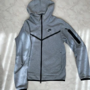 Grå NikeTeck med dragkedja - Nike Tech Fleece hoodiejacka i ljusgrått med svart dragkedja och detaljer. Jackan har huva, lång ärm och en snygg ficka med dragkedja på ärmen. Perfekt för dig som gillar sportig och clean stil. Nike-logga på bröstet och modern passform. Passar även M