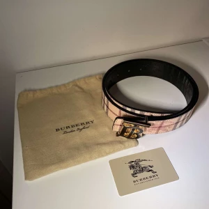 Burberry bälte med klassiskt rutmönster - Säljer ett Burberry bälte i skinn med ikoniskt rutmönster i beige, brunt och svart samt en stor guldfärgad och svart 'TB'-logga som spänne. Bältet är vändbart med ena sidan i klassiskt beige rutigt och andra i svart rutigt. Perfekt accessoar för att lyfta din outfit. 105CM
