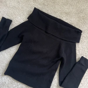 Svart offshoulder stickad tröja - Snygg svart stickad tröja med bred offshoulder-krage. Tröjan har lång ärm och en tightare passform som framhäver siluetten. Perfekt för dig som gillar en stilren och trendig look.
