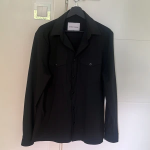 Svart overshirt Shaping New Tomorrow - Stilren svart overshirt från Shaping New Tomorrow med klassisk krage och två bröstfickor med lock och knappar. Skjortan har lång ärm och knäppning framtill, perfekt för en clean och modern look. Nypris 2000kr
