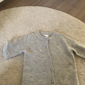 Grå stickad kofta med knappar - Mysig grå stickad kofta med rund hals och knäppning framtill. Perfekt att slänga över en t-shirt eller ha som lager på lager. Klassisk modell med långa ärmar och ribbade muddar för extra komfort. Men saknas en knapp