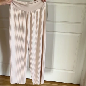 Ljusbeige plisserade vida byxor - Säljer ett par ljusbeige plisserade byxor med hög midja och bred resår. Byxorna har ett flowy och bekvämt wide fit. Materialet känns mjukt och stretchigt, vilket gör dem superbekväma att bära hela dagen.