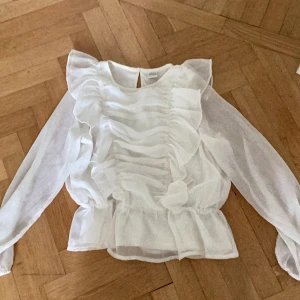 Vit volangblus från Lindex - Säljer en söt vit blus från Lindex med långa ärmar och volangdetaljer längs fram- och baksidan. Blusen är tillverkad i ett lätt och transparent material och har en knapp i nacken. Perfekt för en stilren look!