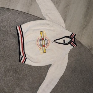 Vit stickad Gant half zip tröja S - Vit stickad tröja från Gant med broderat rowing-märke på bröstet. Half zip med svart, röd och marinblå rand vid krage och muddar. Snygg sportig collegekänsla och ribbade detaljer. Perfekt för dig som gillar preppy stil.