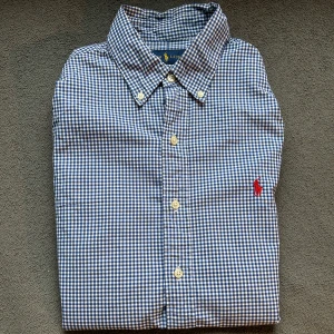 Blårutig Ralph Lauren skjorta - Klassisk blårutig skjorta från Ralph Lauren i bomull med broderad röd logga på bröstet. Storlek M, Cond 7/10 om unte mer, enda "felet" är en liten fläck på framsidan som knappt syns. Hör av er vid frågor och funderingar!
