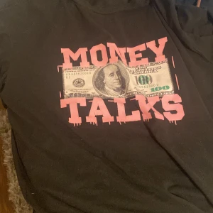 Svart t-shirt med Money Talks-tryck - Svart t-shirt med stort tryck på bröstet som föreställer en hundradollarsedel och texten 'MONEY TALKS' i rosa. T-shirten har klassisk passform och korta ärmar. Materialet känns som bomull och trycket har en streetstyle-vibe.