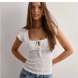 Vit blus med knyt och broderi - Supersöt vit blus med broderade detaljer och knyt framtill. Toppen har korta ärmar och en feminin v-ringning. Perfekt till jeans eller kjol för en fräsch look.