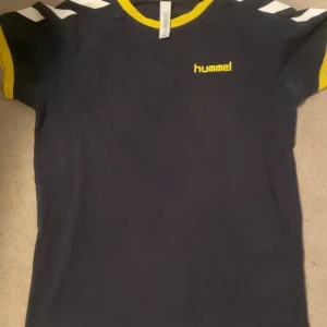 Blå Hummel fotbollströja med gula detaljer - Snygg marinblå fotbollströja från Hummel med gula detaljer och vit logga på bröstet. Klassiska Hummel-pilar på axlarna och stor gul siffra 5 på ryggen. Kortärmad modell med gul kant vid ärmslut och hals. Perfekt för match eller träning.