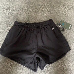 Svarta träningsshorts från Oxide - Svarta träningsshorts från Oxide med elastisk midja och snörning. De är tillverkade i ett lätt och smidigt syntetmaterial som passar perfekt för träning. Klassisk design med diskret logga framtill och kort passform som ger maximal rörelsefrihet. Shortsen har mjuka shorts under också som är perfekt till att träna t.ex springa.