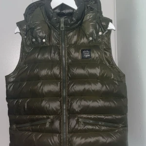 Olivgrön dunväst från Bvaldi - Snygg olivgrön dunväst från Bvaldi med hög krage och avtagbar huva. Västen har två dragkedjefickor framtill och quiltad design. Perfekt att slänga över en hoodie för en chill look. Stängs med dragkedja hela vägen upp.