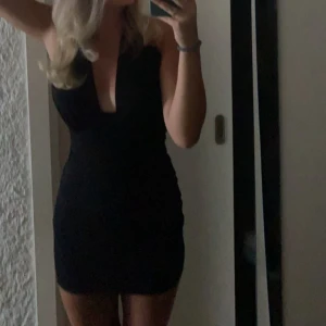 Svart kortklänning med djup v-ringning - Snygg svart kortklänning med djup v-ringning och ärmlös design. Klänningen har en tight passform som framhäver figuren och passar perfekt för dig som vill sticka ut. Enkel men stilren look som är lätt att matcha med accessoarer. Den är halterneck så öppen i ryggen.