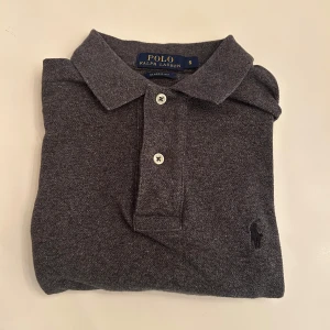 Grå pikétröja Polo Ralph Lauren S - Klassisk grå pikétröja från Polo Ralph Lauren i storlek S. Tröjan har korta ärmar, krage och två knappar framtill. Broderad logga på bröstet och en mjuk bomullskänsla. Perfekt för en clean och stilren look.