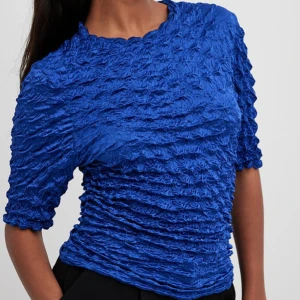 Blå crinkle topp med kort ärm från NA-KD - Säljer en cool blå topp med crinkle-effekt och tight passform från NA-KD. Toppen har korta ärmar och en rund halsringning. Materialet är glansigt och ger en unik struktur som sticker ut. Perfekt för dig som vill ha något annorlunda och trendigt i garderoben.