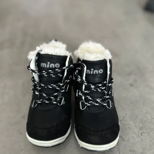 Svarta vinterkängor från Mino - Svarta kängor från Mino med vit fuskpäls runt skaftet och svartvita snören. Skorna har robust sula, metallhål för snörning och detaljer i grått baktill. Perfekta för kalla dagar med sin mysiga insida och snygga kontraster.