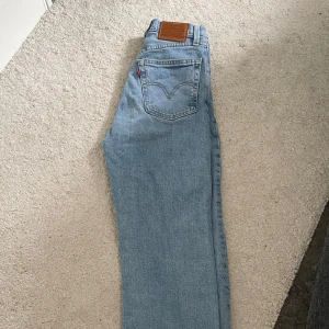 Levi's Ljusblåa jeans straight fit - Klassiska Levi's jeans i ljusblå denim med raka ben och hög midja. Jeansen har fem fickor, knappgylf och den ikoniska läderpatchen bak i midjan. Perfekta för en avslappnad och tidlös stil. Midja 24, längd 28. De är lite insydda i midjan.