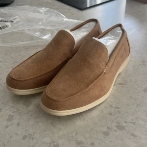 Helt nya mockaloafers  - Snygga loafers i beige mocka med stilren design och vit platt sula. Loafersen är aldrig använda. Perfekta för dig som gillar en clean och avslappnad look.