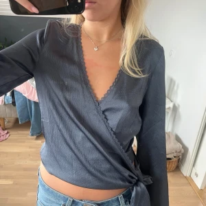 Mörkgrå omlottblus med knyt - Supersnygg mörkblå omlottblus med v-ringning och knyt i sidan. Blusen har långa, vida ärmar och fina spetsdetaljer längs kanterna. Säljer pgd att jag inte får använding av den tyvärr!💓🥰