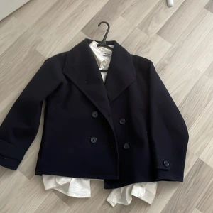 Svart skepparkavaj från Gina - Stilren svart skepparkavaj från Gina med dubbelknäppning och bred krage. Jackan har en klassisk, boxig passform och är tillverkad i ett tjockare material som passar perfekt till kyligare dagar. Snygg att slänga över en skjorta eller hoodie . 