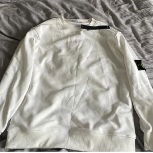 Vit sweatshirt från Stone Island - Vit långärmad sweatshirt från Stone Island med klassisk rund hals och ribbade muddar. Tröjan har den ikoniska loggan på vänster ärm och är gjord i mjuk bomull. Perfekt för en clean och stilren look.