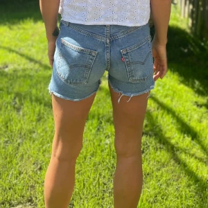  Levi's ljusblå jeansshorts med frans - 2 par klassiska ljusblå jeansshorts från Levi's med råa fransade kanter och femficksmodell. De har en normal passform och tydliga Levi's-detaljer som läderpatch och röd etikett på bakfickan. Perfekta för sommardagar och enkel att matcha med allt. Säljs för 130kr st. 