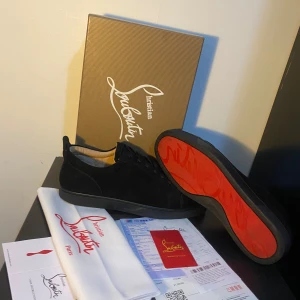  Louboutin skor - Svarta sneakers från Christian Louboutin i mocka med ikonisk röd sula och diskret snörning. Snygg och stilren design med rund tå och platt sula. Perfekta för dig som vill sticka ut med exklusiva detaljer och lyxig känsla.