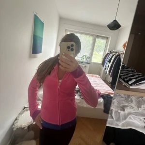 Rosa hoodie från juicy - Säljer en rosa hoodie från juicy med dragkedja framtill och huva. Jackan har lila muddar och blå kant nertill, samt två fickor. Perfekt för dig som gillar sportig och färgglad stil.