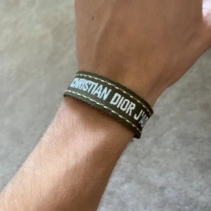 Christian Dior J'Adior armband - Snyggt armband från Christian Dior i militärgrön färg med vit broderad text 'CHRISTIAN DIOR J'ADIOR'. Armbandet är gjort i vävd textil och har fransar i ändarna för en cool och avslappnad vibe.