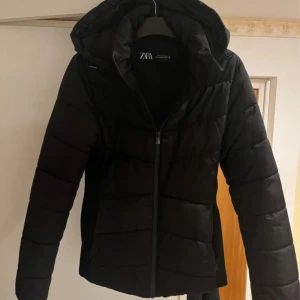 Svart pufferjacka från Zara - Säljer en svart pufferjacka från Zara med huva och dragkedja framtill. Jackan är vadderad och har en klassisk, enkel design som passar perfekt när det är kallt ute. Snygg och stilren modell som är lätt att matcha med olika outfits.