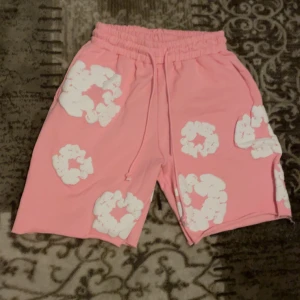 Rosa shorts från Denim Tears S - Säljer ett par rosa shorts från Denim Tears med vita blommiga tryck över hela plagget. De har elastisk midja med snörning och är tillverkade i mjuk bomull. Perfekta för en chill sommarlook med unik vibe.