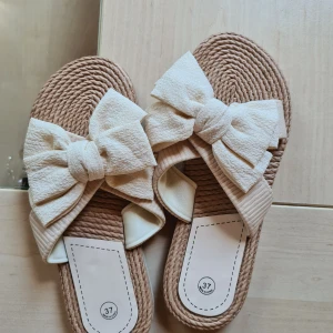 Beige sandaler med stor rosett - Snygga sandaler i beige med flätad sula och bred rem i tyg, dekorerad med en stor, mjuk rosett framtill. Perfekta för sommardagar och ger en söt touch till din outfit. Platt sula och öppen tå för extra komfort.