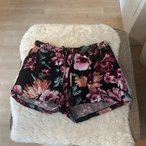 Blommiga svarta shorts med snörning - Snygga svarta shorts med stort blommönster i rosa, vitt och grönt. De har en bekväm midja med snörning och lös passform, perfekta för varma dagar. Lätta och luftiga, passar perfekt till sommaren. Eller att sova i