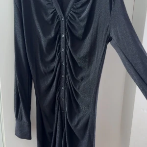 Svart glittrig långärmad klänning - Snygg svart klänning med glittrig finish och långa ärmar. Klänningen har knappar hela vägen framtill, markerade manschetter och coola rynkade detaljer längs bröst och midja. Materialet är stretchigt med metallictråd för extra shine.