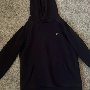 Svart hoodie från GANT med logga - Svart hoodie från GANT med liten broderad logga på bröstet. Klassisk modell med huva och känguruficka framtill. Perfekt basic-plagg med stilren look och skön passform.