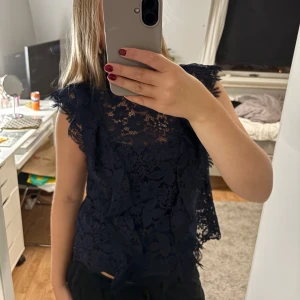 Mörkblå spetsblus med volang - Supersnygg mörkblå blus från Zara i spets med volangdetaljer och ärmlös design. Blusen har ett feminint blommönster i spetsen och hög hals. Perfekt för dig som vill ha en elegant och trendig look.